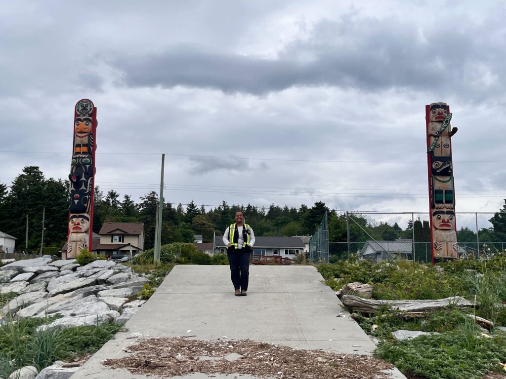 An engineer stands between two totem poles on the beach in Tla'amin Nation.